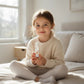 Young girl sitting on a bed holding a bottle in a bright room.