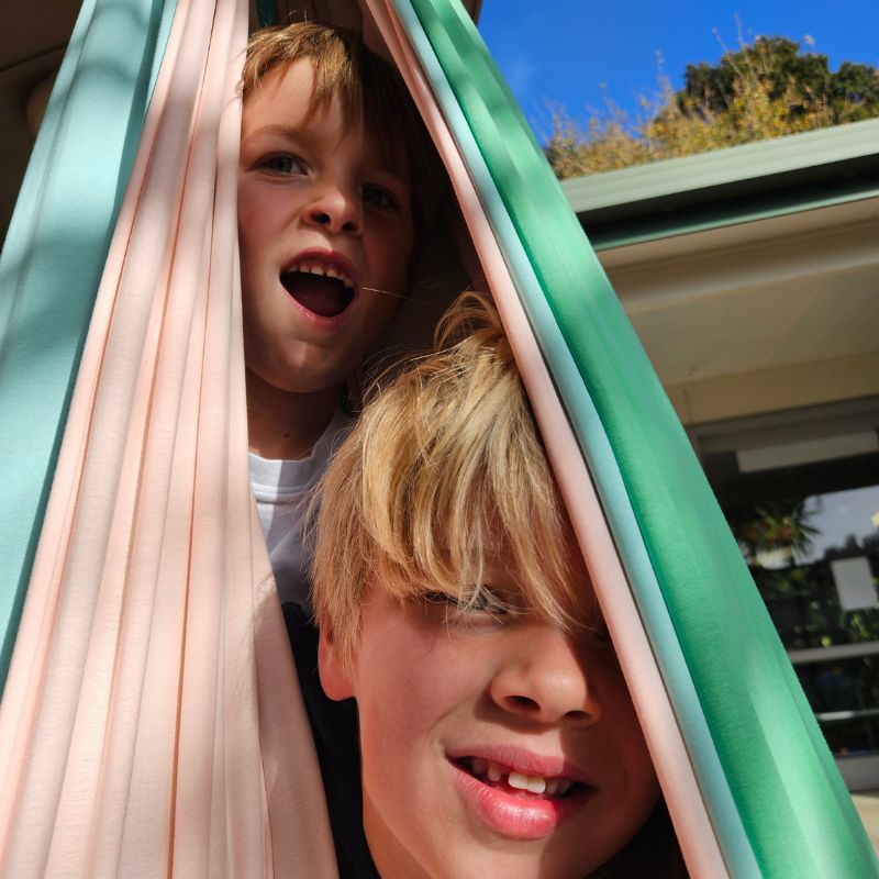 Two children peeking from behind colorful fabric with a building and trees in the background.