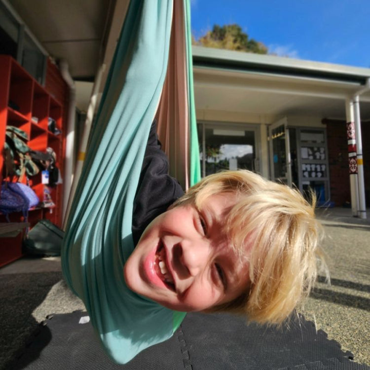 Child hanging upside down in a green fabric swing with a school building in the background