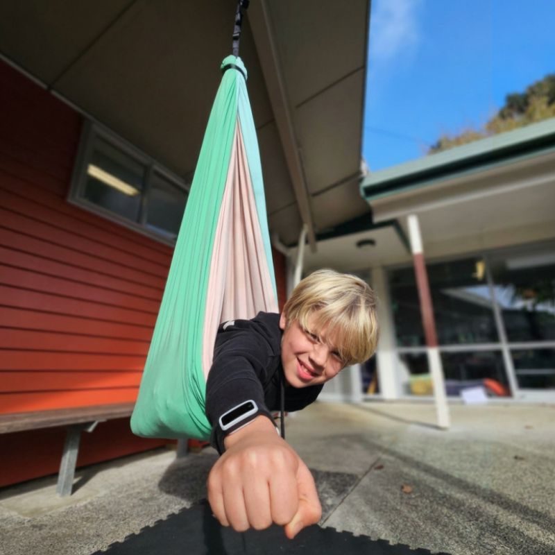 Person hanging from a green and pink hammock outside a building with a clear sky.