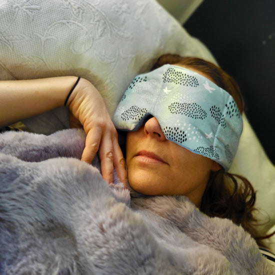 A person lying down wearing a blue weighted sleep mask with a pattern of dots and lines, over a white background.