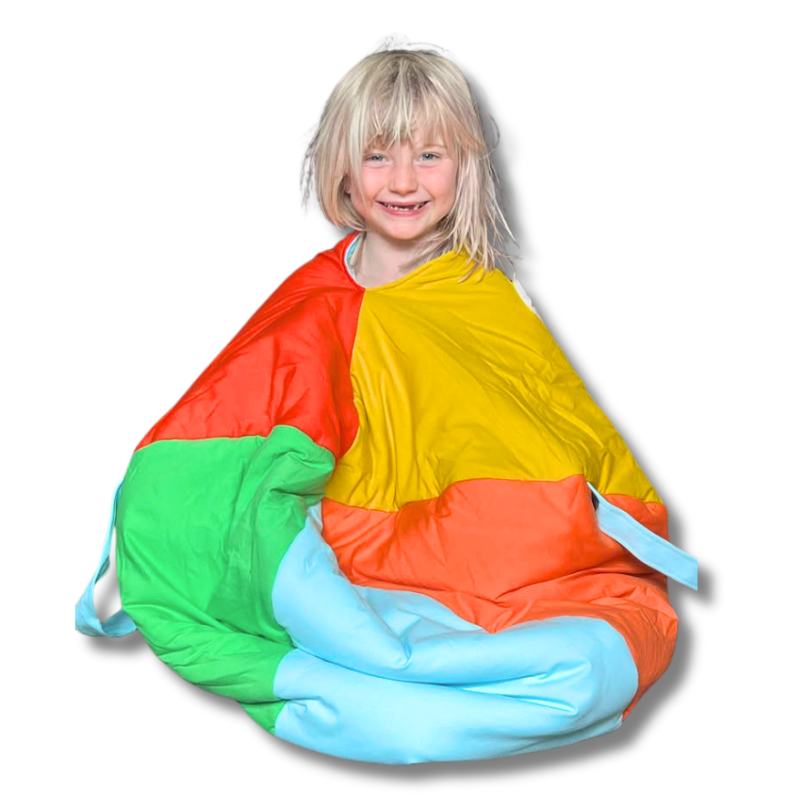 A child sitting cross-legged while inside a multicolored hideaway sack with openings for arms and legs, made of soft fabric.