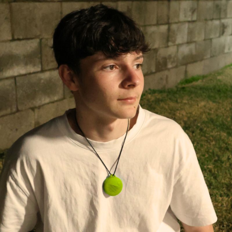 Person wearing a white shirt and a necklace with a green pendant, sitting against a brick wall.