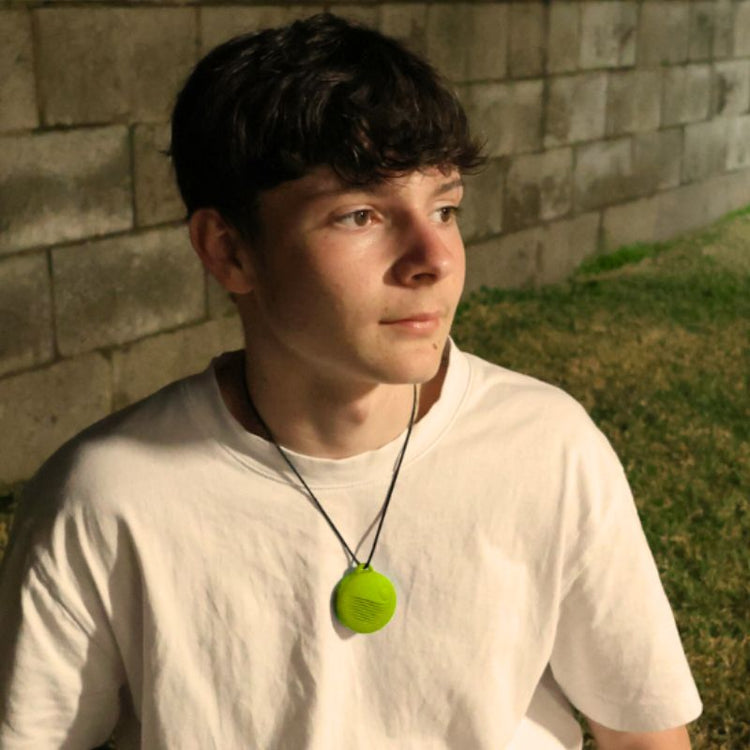 Person wearing a white shirt and a necklace with a green pendant, sitting against a brick wall.