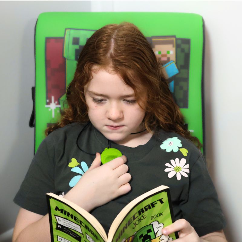 Child reading a Minecraft joke book with a green cushion featuring Minecraft characters in the background.