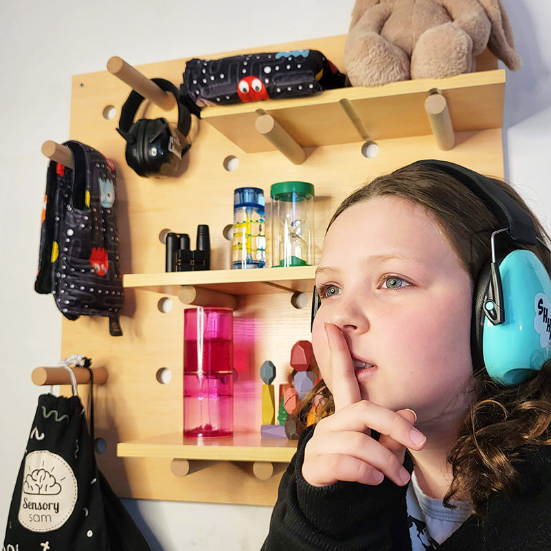 A child sitting in front of a wooden peg board with various sensory toys and equipment arranged on it, including bottles, plush toys, and sensory tools.