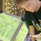 A child looking at a green illuminated LED infinity cube with a remote control in hand, set against a blurred background of lights.