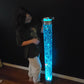 A person standing next to a tall cylindrical LED bubble tube lamp that is lit up with blue bubbles visible inside it.