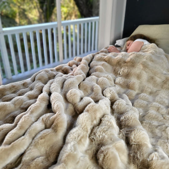 A child appears to be sleeping soundly under a beige weighted blanket with a bumpy fur texture, with a balcony visible in the background.