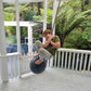 Child on a large black ball swing on a porch with greenery in the background