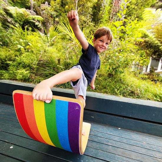 A child standing on a balance board with a rainbow felt grip, smiling and raising one arm.