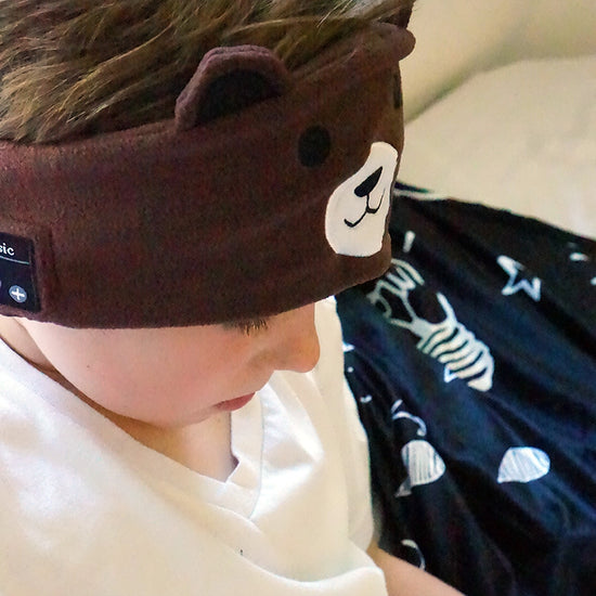 A child wearing a brown pair of ear muffs with a bear design on the top, connected to a device via a 3.5 mm cable.