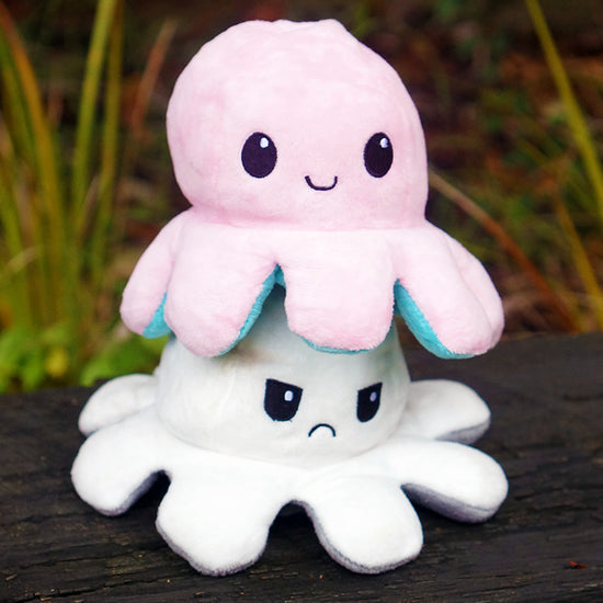 A plush toy resembling an octopus with reversible facial expressions, one side happy and one side sad, set against a natural background.