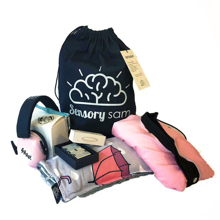 A collection of sensory stimulation products including a pink body sock, a lap pad, shh muffs, a wooden fidget roller, and a flippy fidget rad roller, all displayed in front of a denim sack with the logo &