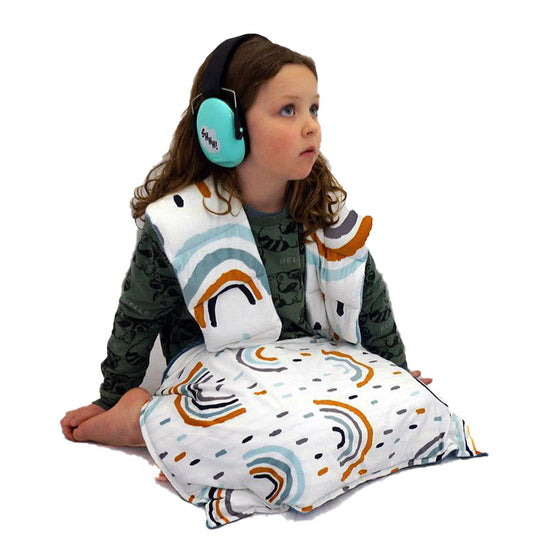 A child wearing headphones and a rainbow-patterned sensory sack with shoulder huggers and a lap pad.