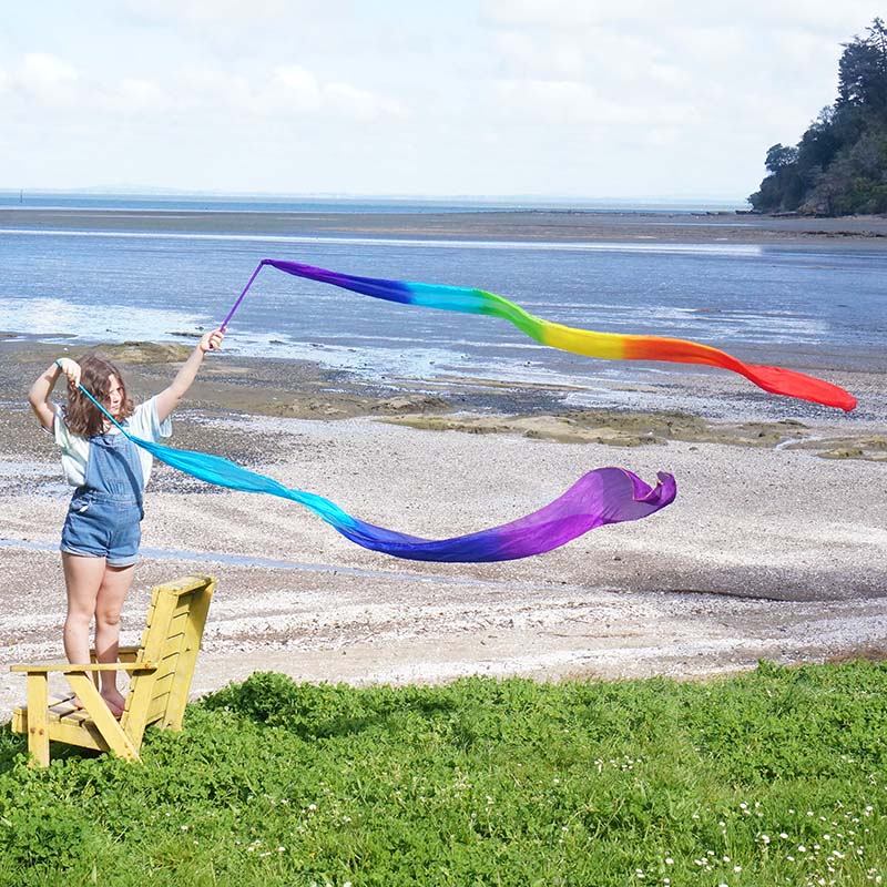A person standing on a grassy area by the beach is holding up a long, colorful silk ribbon that is swirling in the wind.
