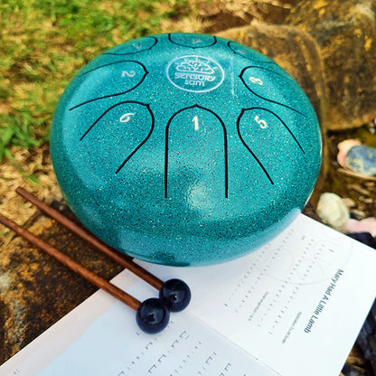 A steel tongue drum with 8 keys, displayed alongside two rubber mallets, a carry case, and a song book. The drum has a metallic turquoise finish and is placed on a natural stone setting.