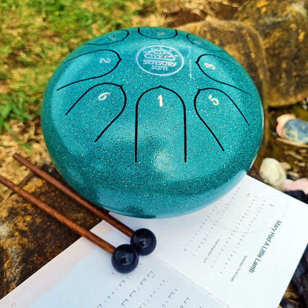 A steel tongue drum with 8 keys, displayed alongside two rubber mallets, a carry case, and a song book. The drum has a metallic turquoise finish and is placed on a natural stone setting.