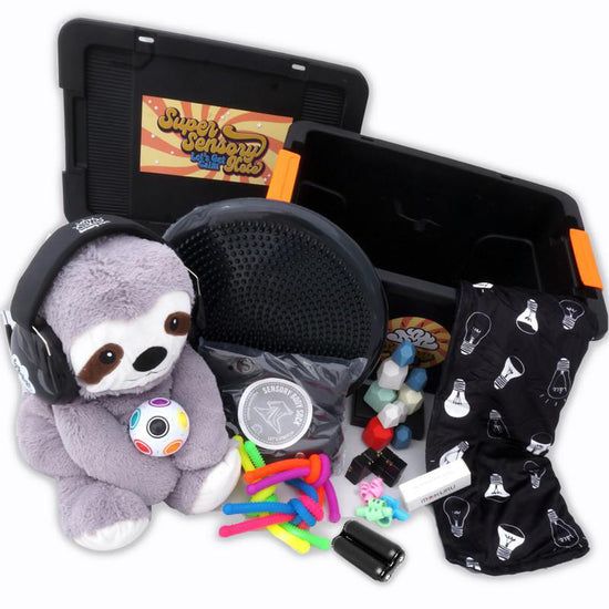 A collection of sensory toys and accessories including a weighted lap pad, a sloth plush toy, a black wobble cushion, shh muffs, a balance stone, a fidget, a puzzle ball, pencil grips, and a variety of strings presented alongside a heavy-duty storage box.