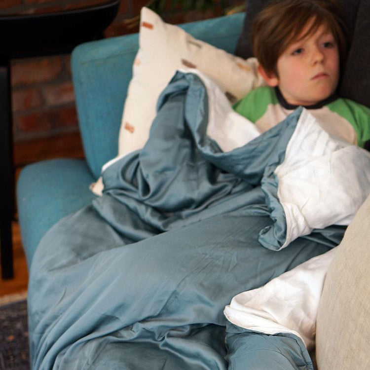 A child appears to be resting or sleeping on a couch with a blue weighted blanket covering them, with a white pillow beside them.
