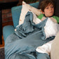 A child appears to be resting or sleeping on a couch with a blue weighted blanket covering them, with a white pillow beside them.